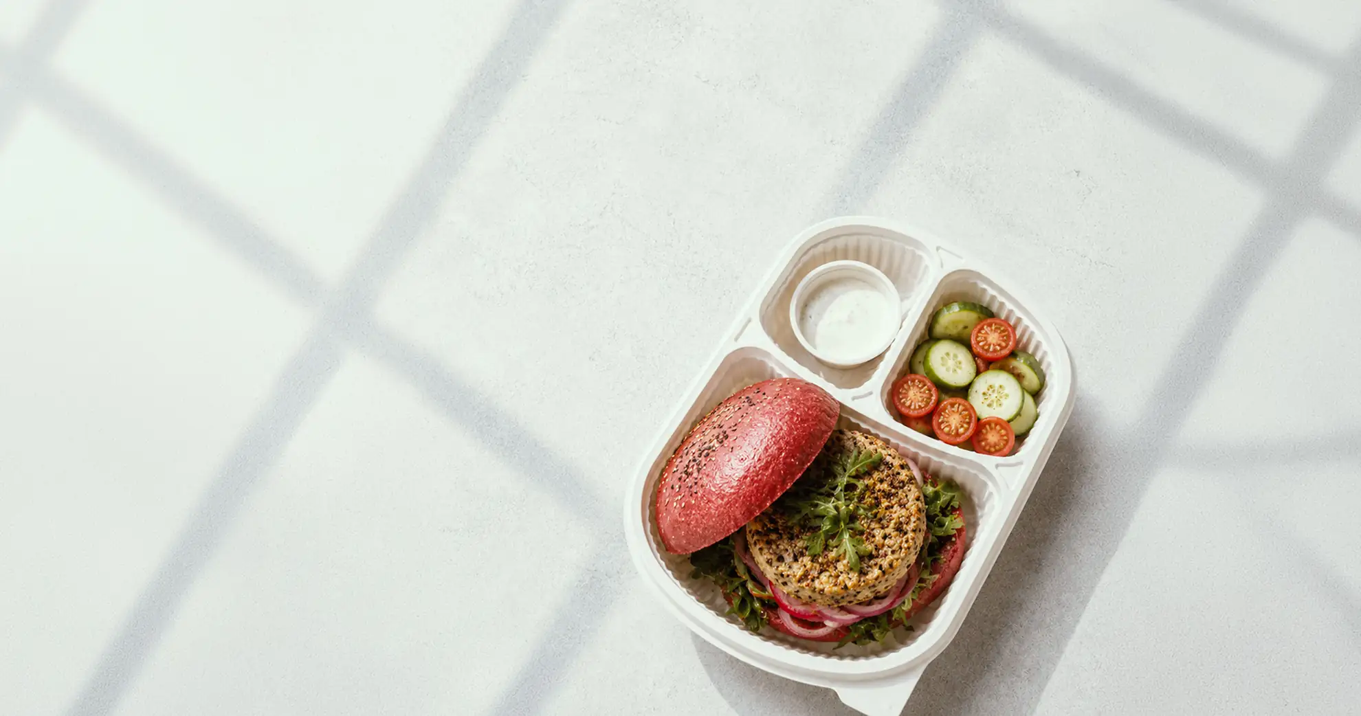 Gourmet lunch box with a quinoa-crusted burger on a beetroot bun, cucumber and cherry tomato salad and a creamy dip, for premium office catering in Bangalore